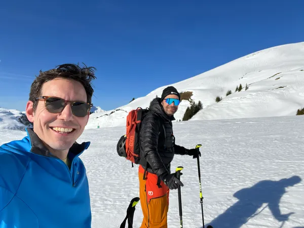 Selfie on a sunny ski tour: Christoph in front, Markus behind him with poles and a red backpack, snowy slopes under a blue sky